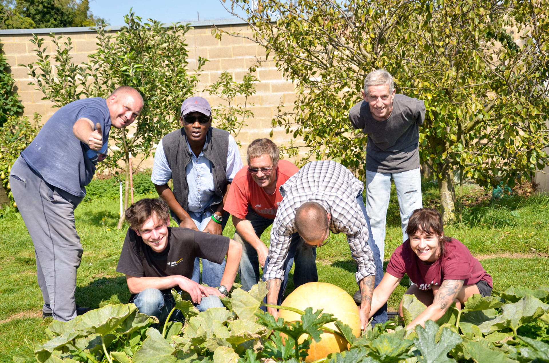 Group of six people outdoors smiling and posing around a large pumpkin growing in a garden.