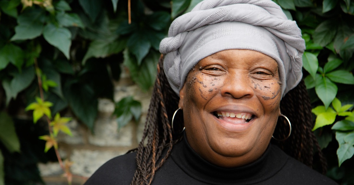 Smiling woman with face tattoos, wearing a light gray headwrap and hoop earrings, standing in front of green leafy wall.