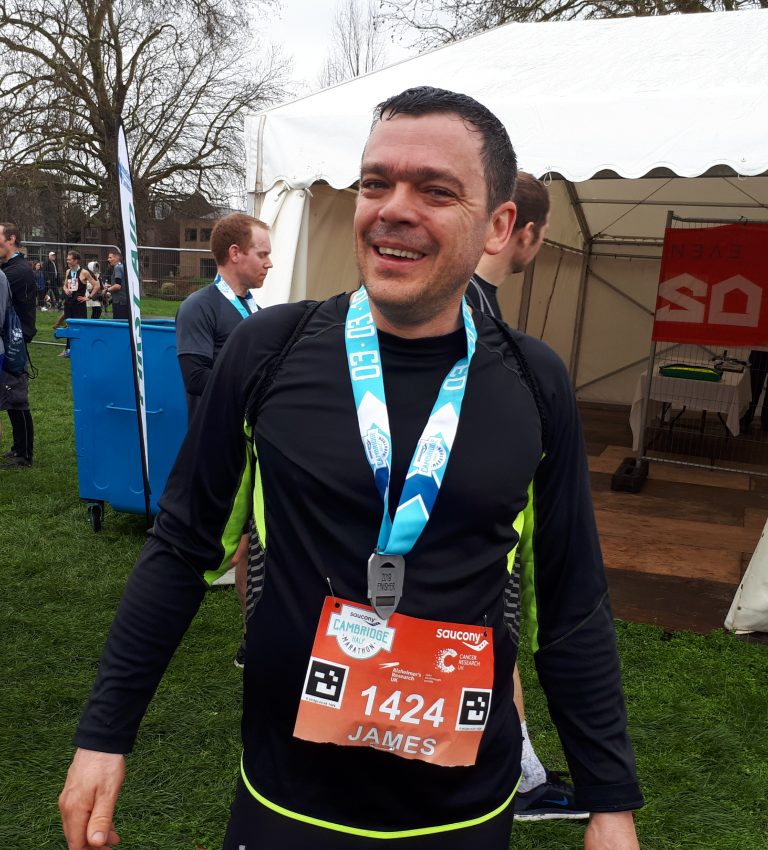 Smiling man wearing a black and neon green athletic shirt with a blue race finisher medal and race bib number 1424 labeled James standing outdoors near a white tent.