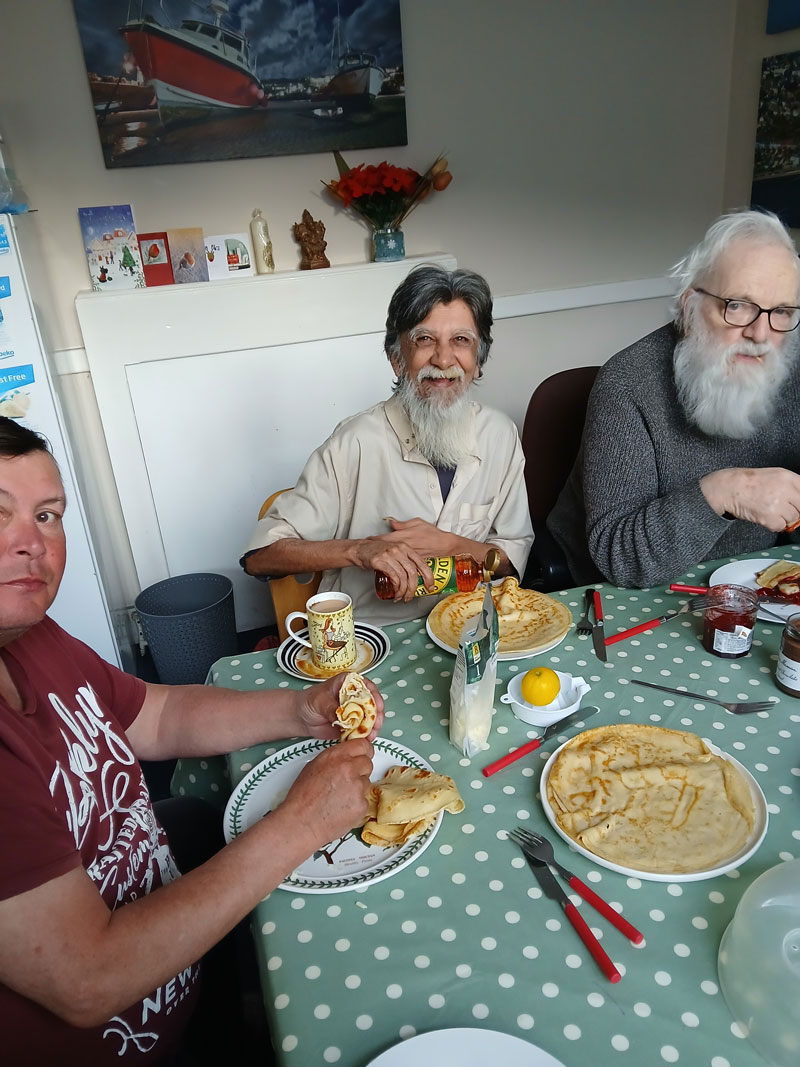 Three men sitting around a polka dot tablecloth at a table, eating crepes and drinking tea, one man pouring syrup.