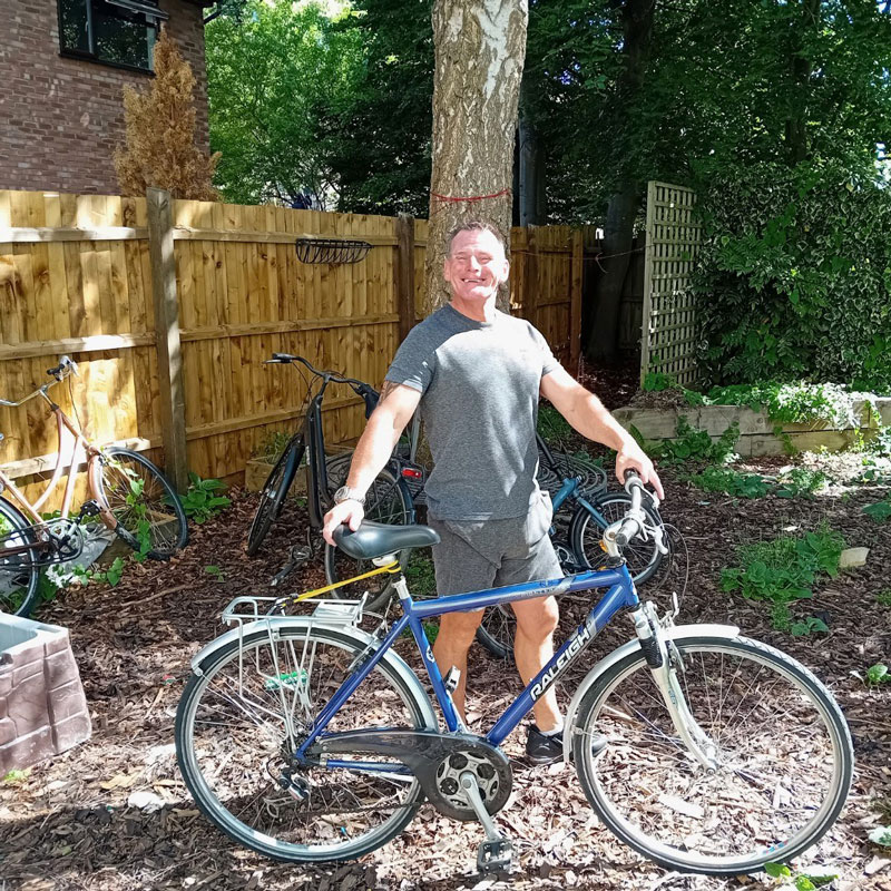 Smiling man in gray shorts and t-shirt standing outdoors holding a blue Raleigh bicycle in a garden area with trees and wooden fence.