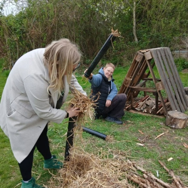 Two people outdoors handling dried straw and black tubes near a wooden pallet structure on grass.