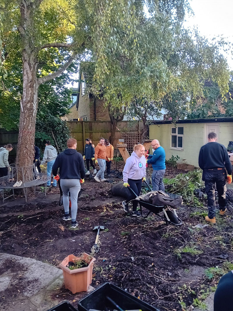 Group of people working together to clear and clean a garden area with trees, gardening tools, and a wheelbarrow.