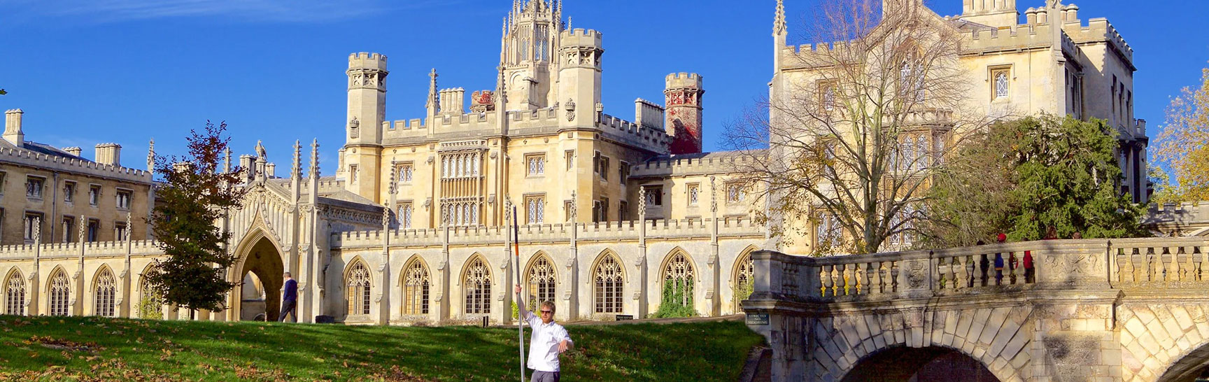 Historic stone building in Cambridge with arched windows and battlements beside a grassy area and a stone bridge under a clear blue sky.