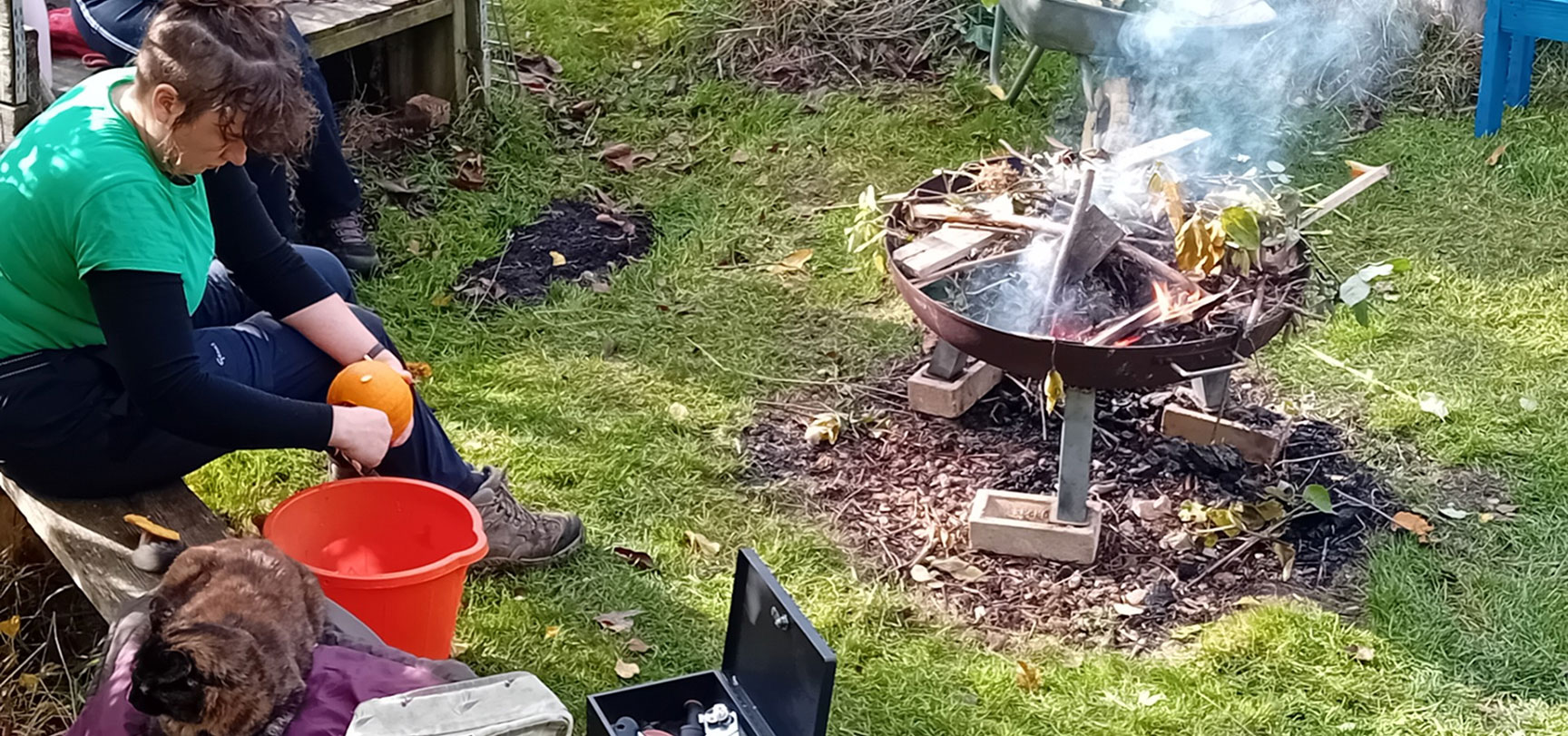 Person sitting on a wooden bench outdoors carving a small pumpkin near a smoking fire pit made of metal.