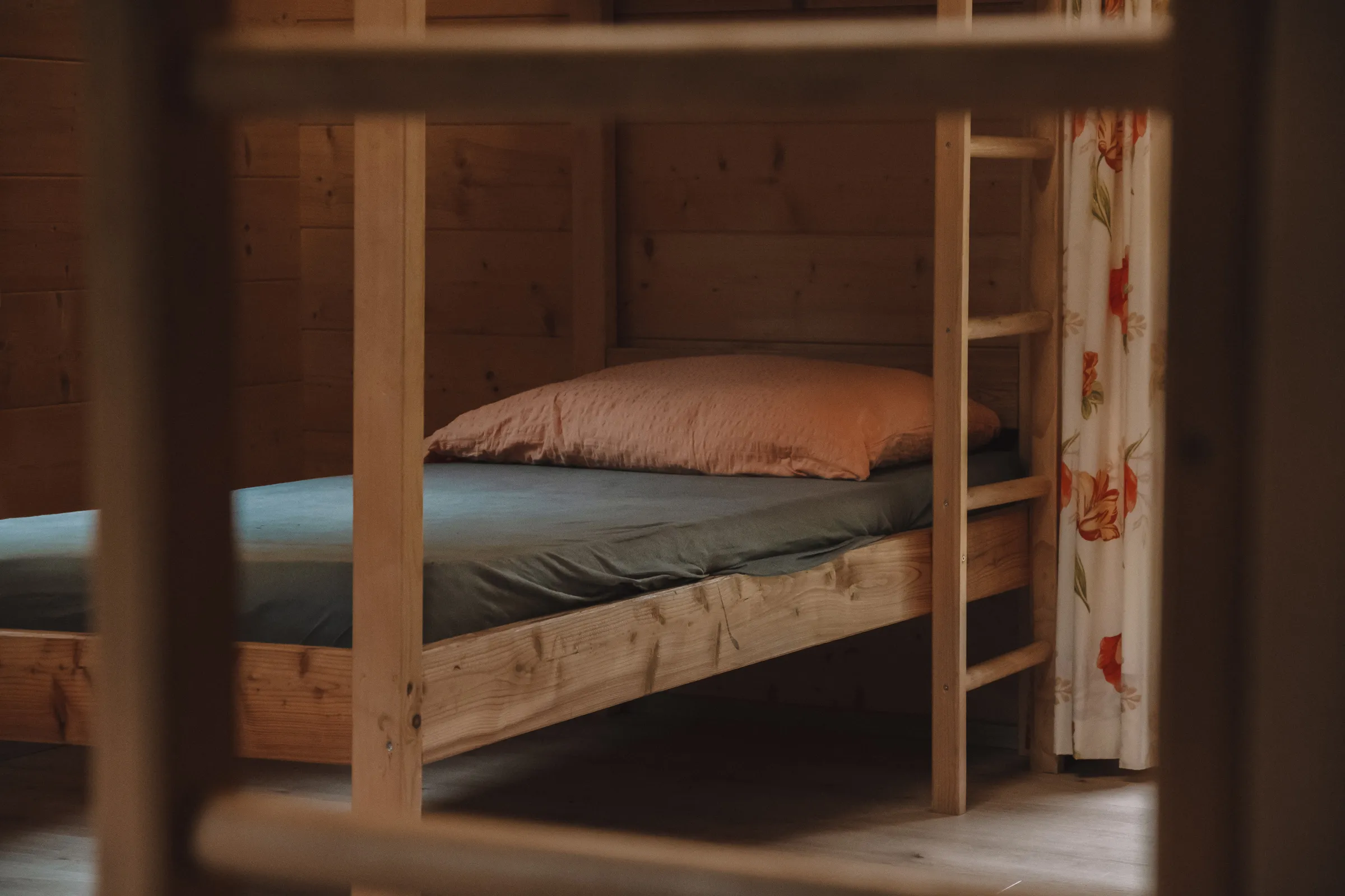 Holz-Etagenbett mit blauem Bettlaken und oranger Kopfkissen in einer gemütlichen Hütte mit Holzvertäfelung und blumengemustertem Vorhang.
