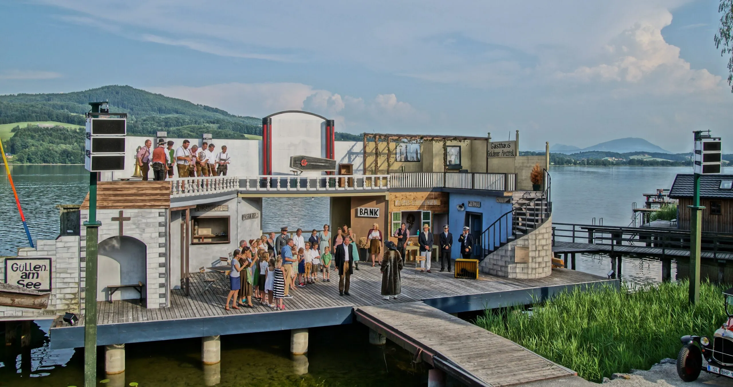Freiluftbühne mit einer Theatergruppe, die eine Szene vor einem Gebäudeensemble mit Schildern wie 'BANK', 'POLIZEI' und 'Güllen am See' aufführt, im Hintergrund ein See und grüne Hügel.