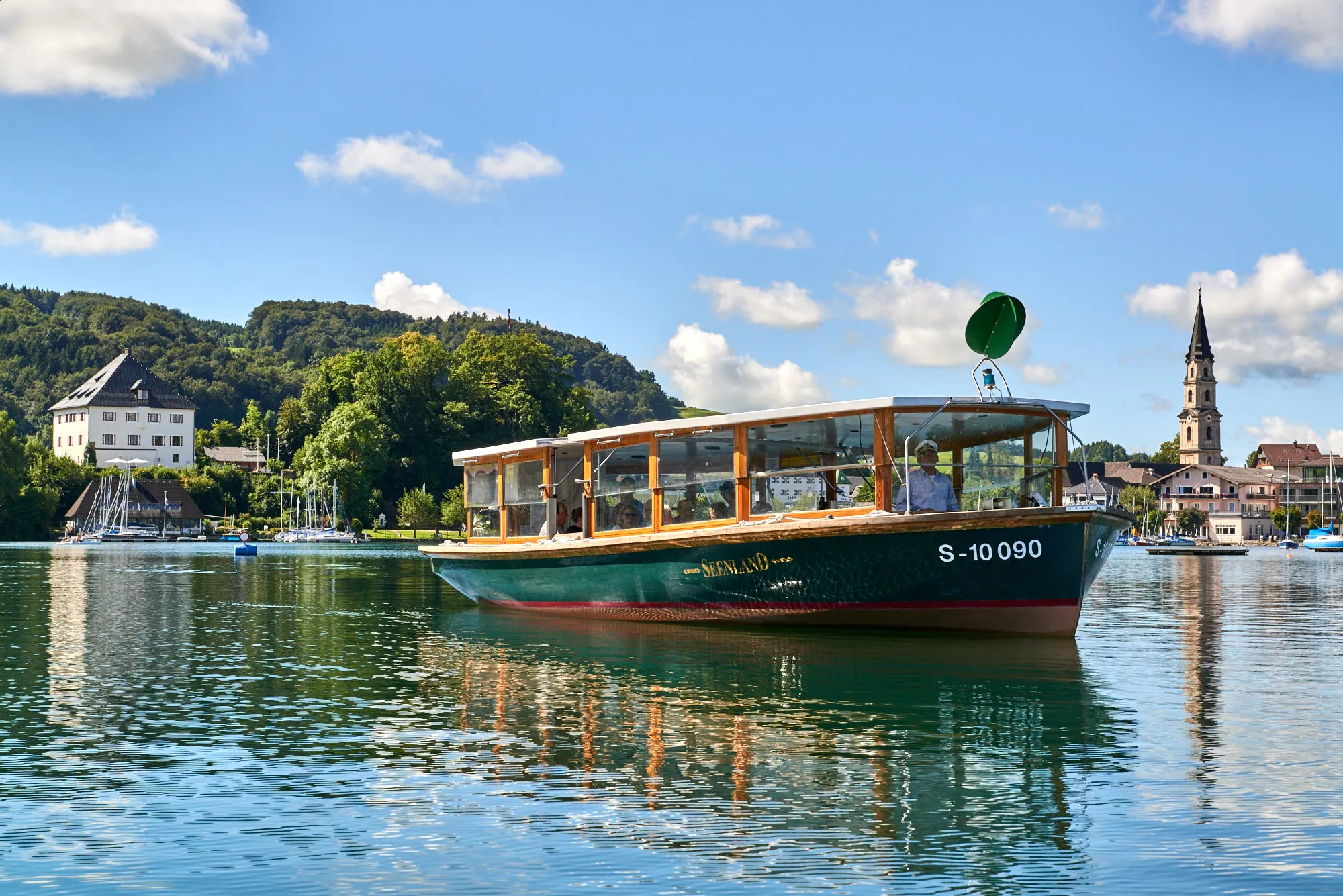 Ein grünes Passagierboot mit der Aufschrift 'SEENLAND' fährt auf einem ruhigen See, umgeben von grünen Hügeln, Segelbooten und Gebäuden, darunter ein weißes Schloss links und ein Kirchturm rechts, unter einem blauen Himmel mit wenigen Wolken.