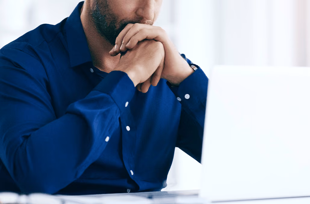 Um homem barbudo, vestindo uma camisa social azul, sentado diante de um laptop com as mãos cruzadas perto da boca.