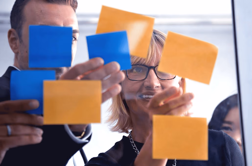Um grupo de colegas organizando post-its coloridos em tons de azul e laranja em uma parede de vidro transparente durante uma sessão de brainstorming.