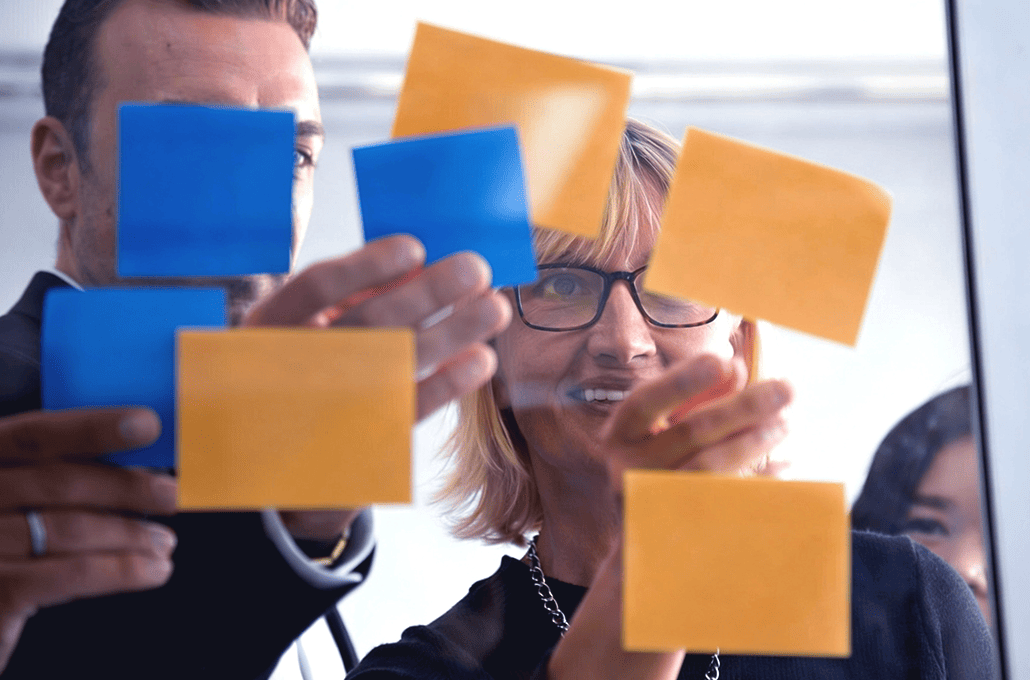 A group of colleagues arranging colorful blue and orange sticky notes on a transparent glass wall during a brainstorming session.
