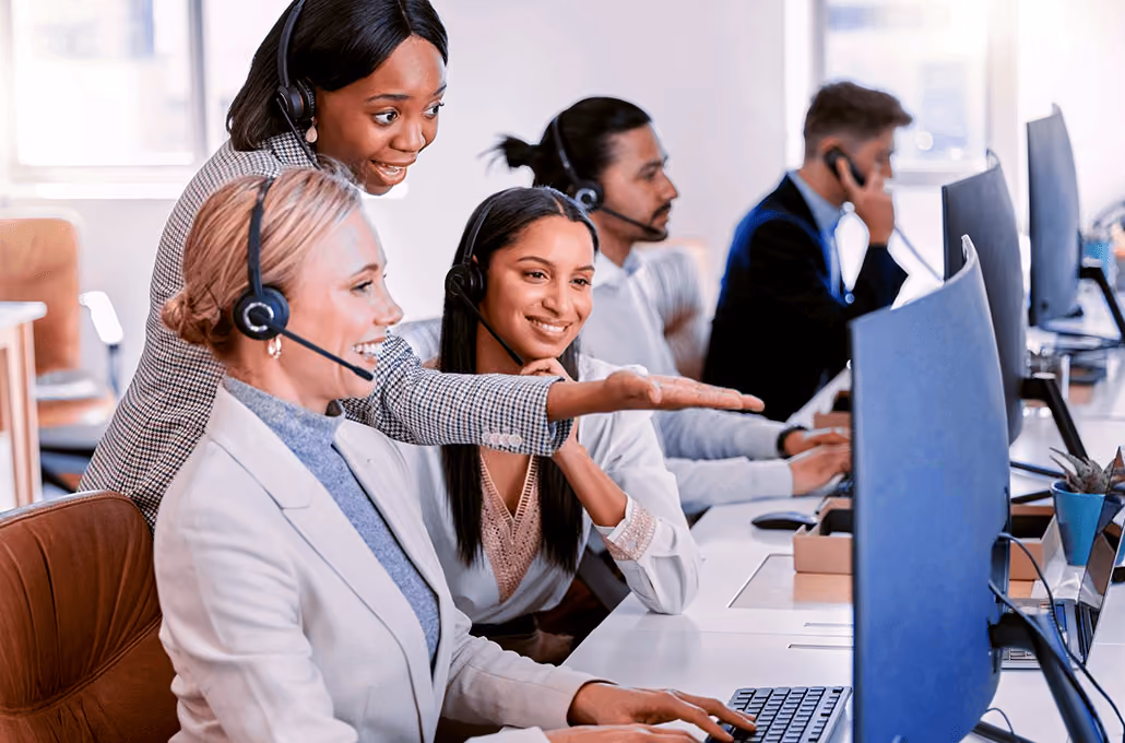 An office setting showing customer service agents wearing headsets, with two women smiling and pointing at a large computer monitor.