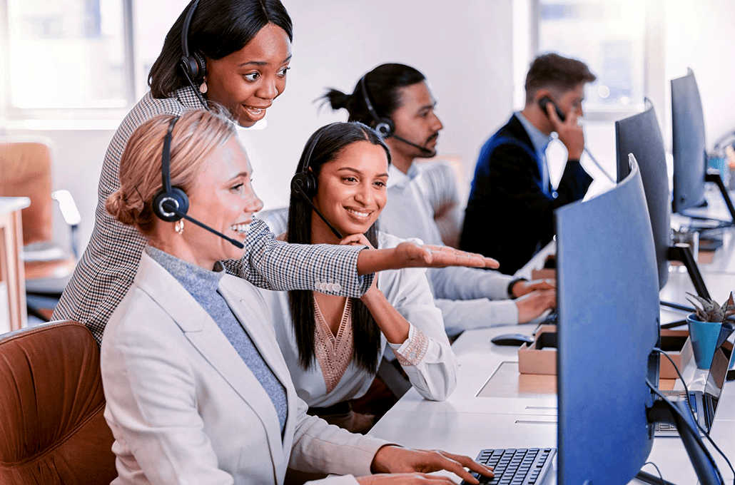 An office setting showing customer service agents wearing headsets, with two women smiling and pointing at a large computer monitor.