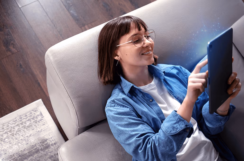 A smiling woman with glasses and dark hair, comfortably leaning back on a grey sofa while using a digital tablet.