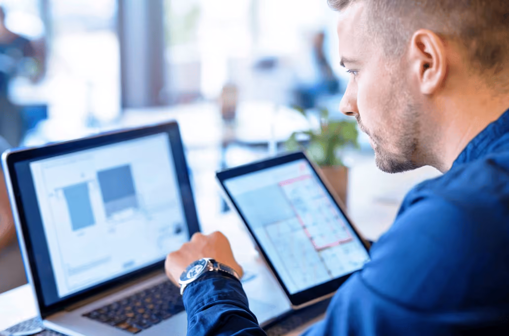Um homem de camisa azul olhando atentamente para duas telas em sua mesa: um laptop exibindo gráficos de dados.