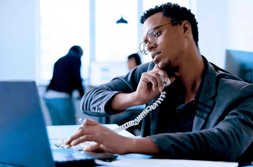 A focused professional wearing glasses talks on a landline phone while working at his desk on a laptop in a modern office environment.