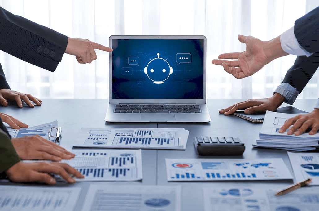 A team of professionals sits around a conference table covered in financial reports and charts, pointing toward a laptop displaying a glowing AI chatbot icon on its screen.
