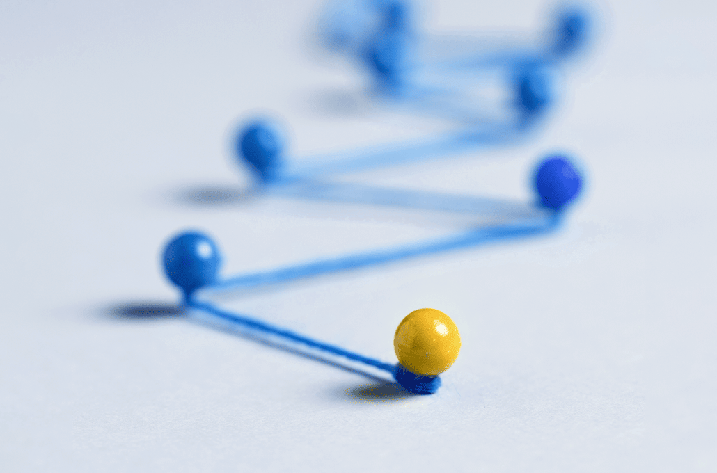A minimalist close-up shot showing blue pushpins connected by a zigzagging blue string across a white surface, leading to a single bright yellow pin that stands out at the front.