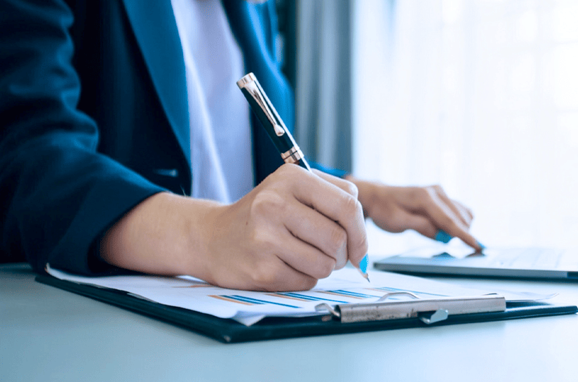A professional wearing a dark blazer, holding a pen to write on documents attached to a clipboard while using a laptop trackpad in a brightly lit office setting.