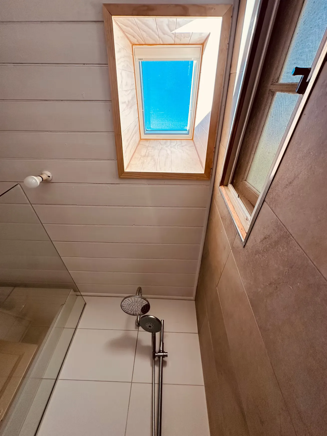 Shower area with a rain showerhead, white tiled wall, brown tiled wall, wooden ceiling, and a rectangular skylight showing blue sky.