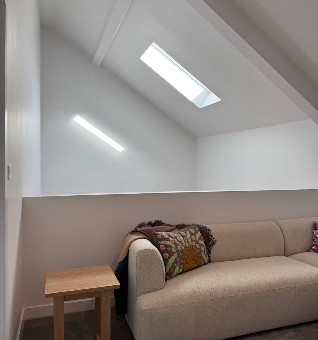 Minimalist living room with beige sofa, floral pillow, wooden side table, and skylight windows in slanted ceiling.