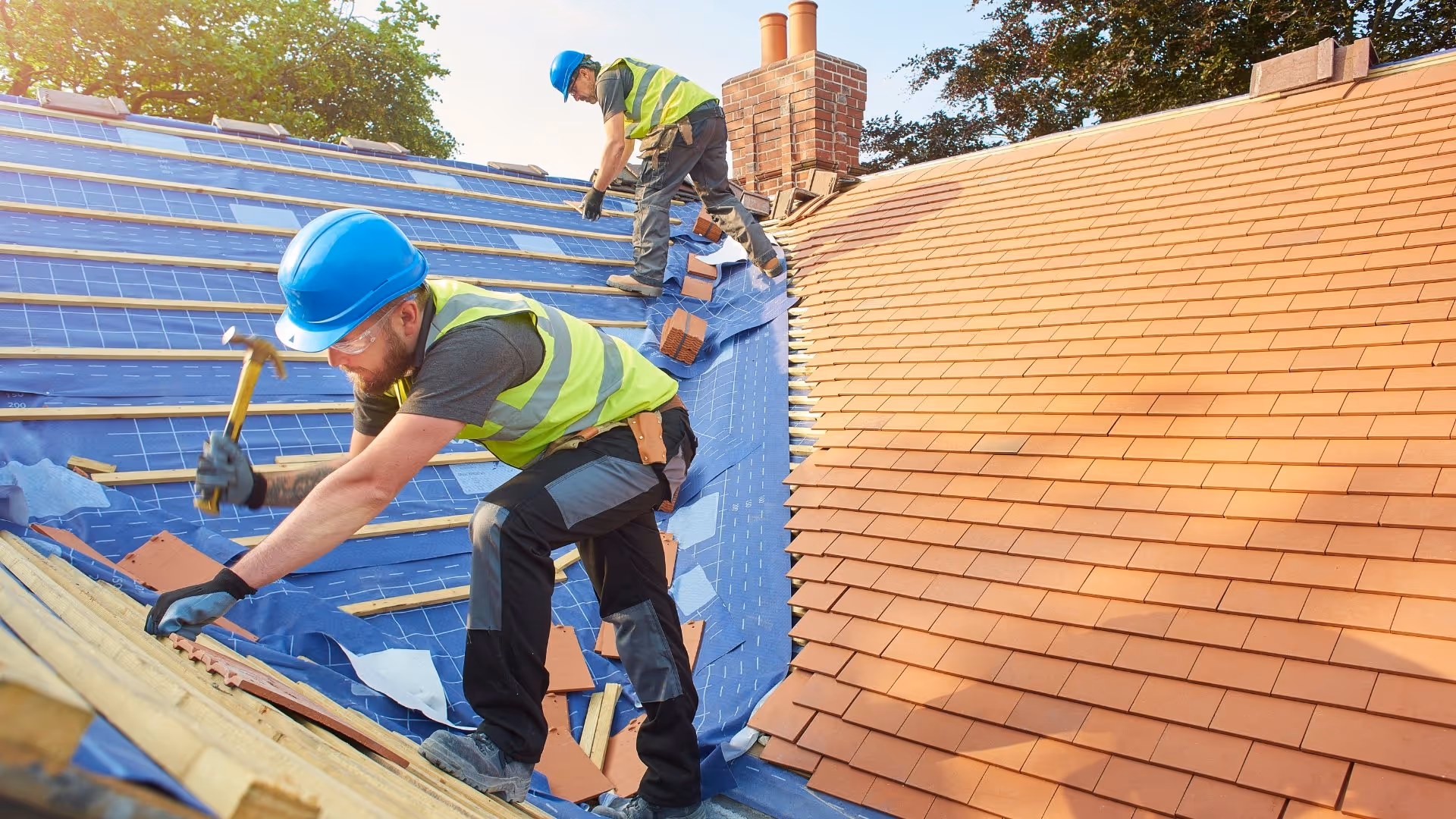 Two construction workers wearing blue helmets and yellow vests installing roof tiles on a house under a clear sky.