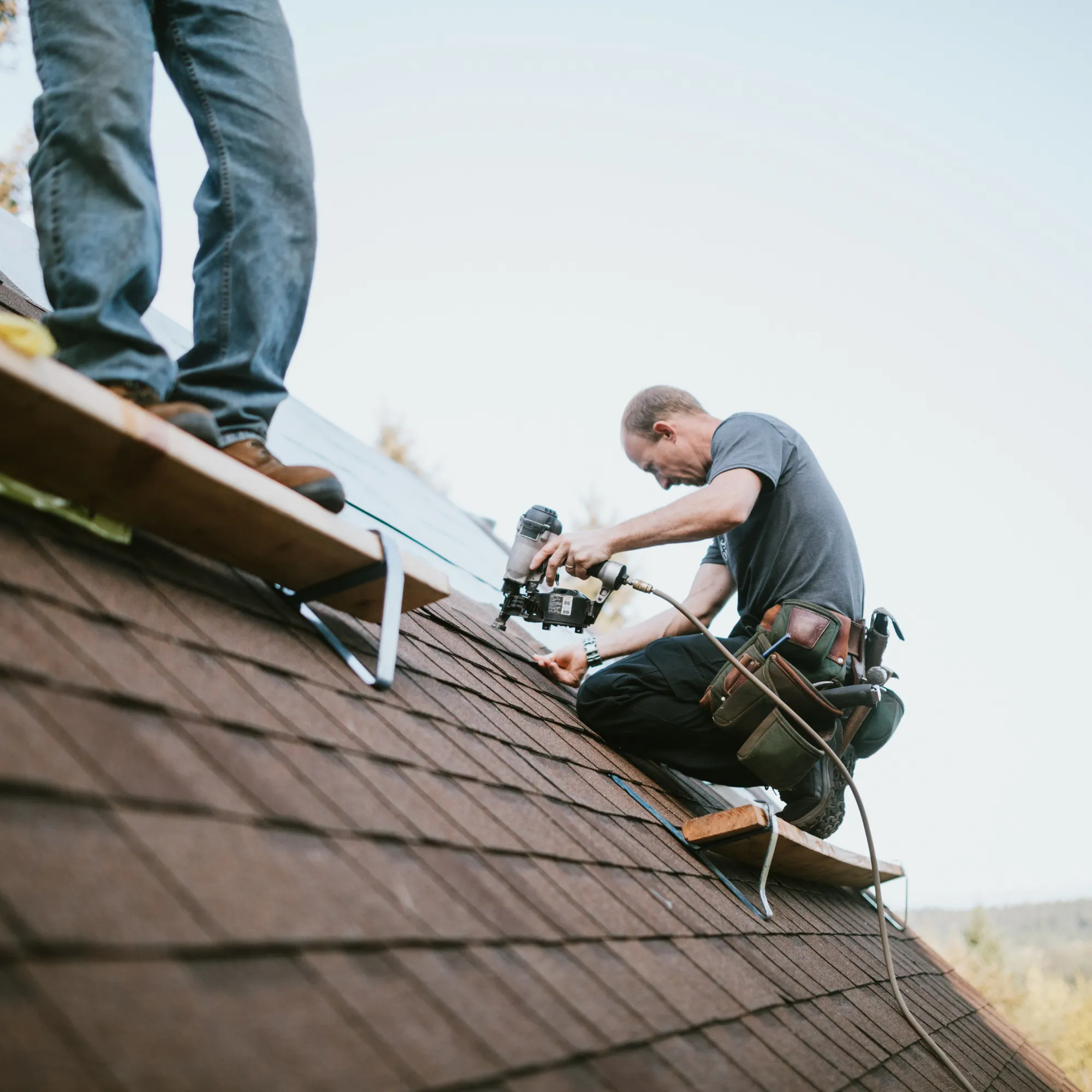 Construction worker kneeling on a roof using a nail gun with another person standing nearby on a wooden plank.