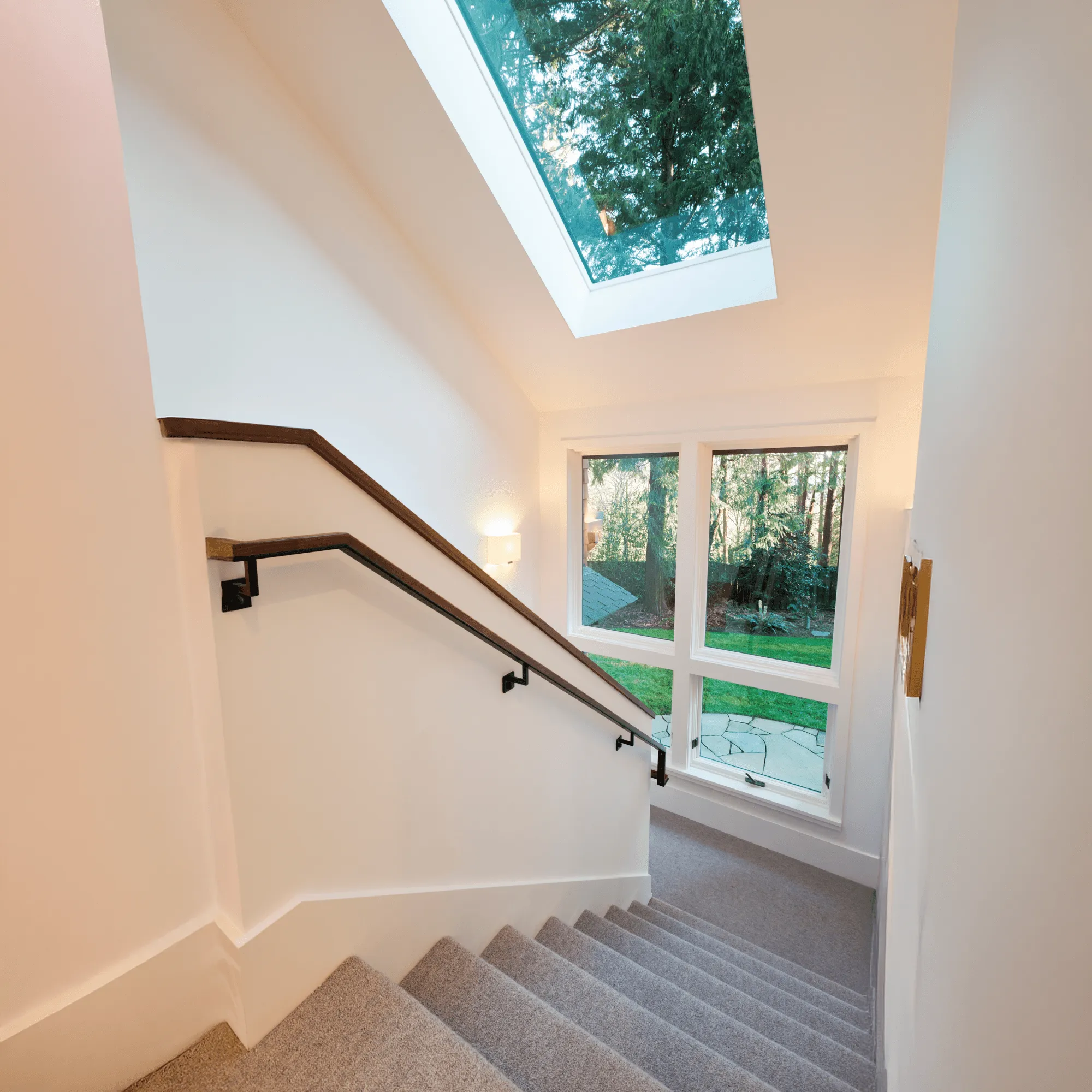 Indoor staircase with grey carpet, wooden handrail, skylight above, and large window overlooking a green garden.