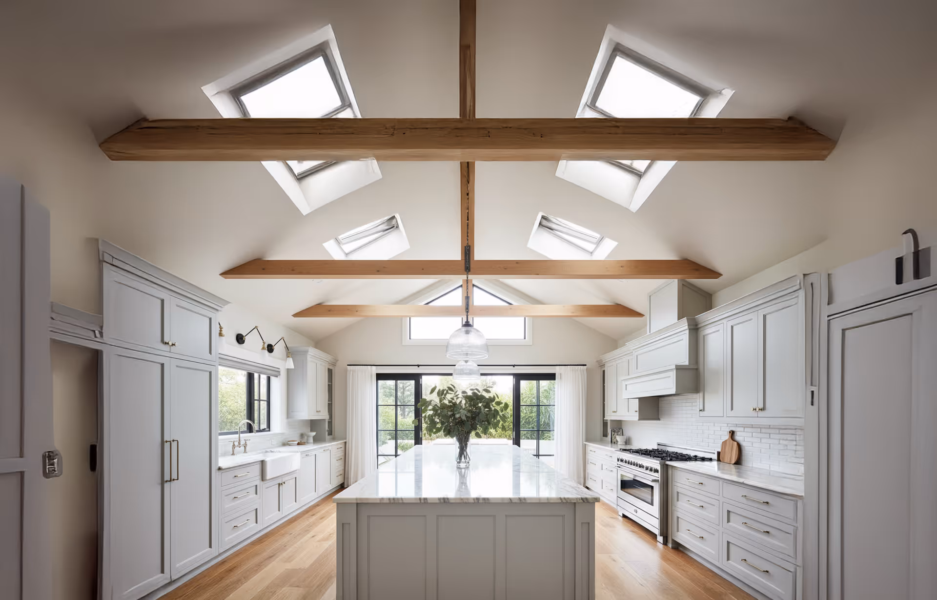 Bright modern kitchen with large marble island, wooden beams, skylights, and black-framed glass doors.
