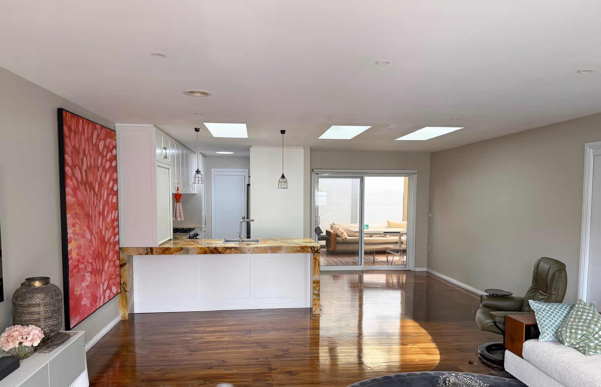 Modern open kitchen with white cabinets, marble countertop, two pendant lights, hardwood floor, and adjacent living area with couch and armchair.