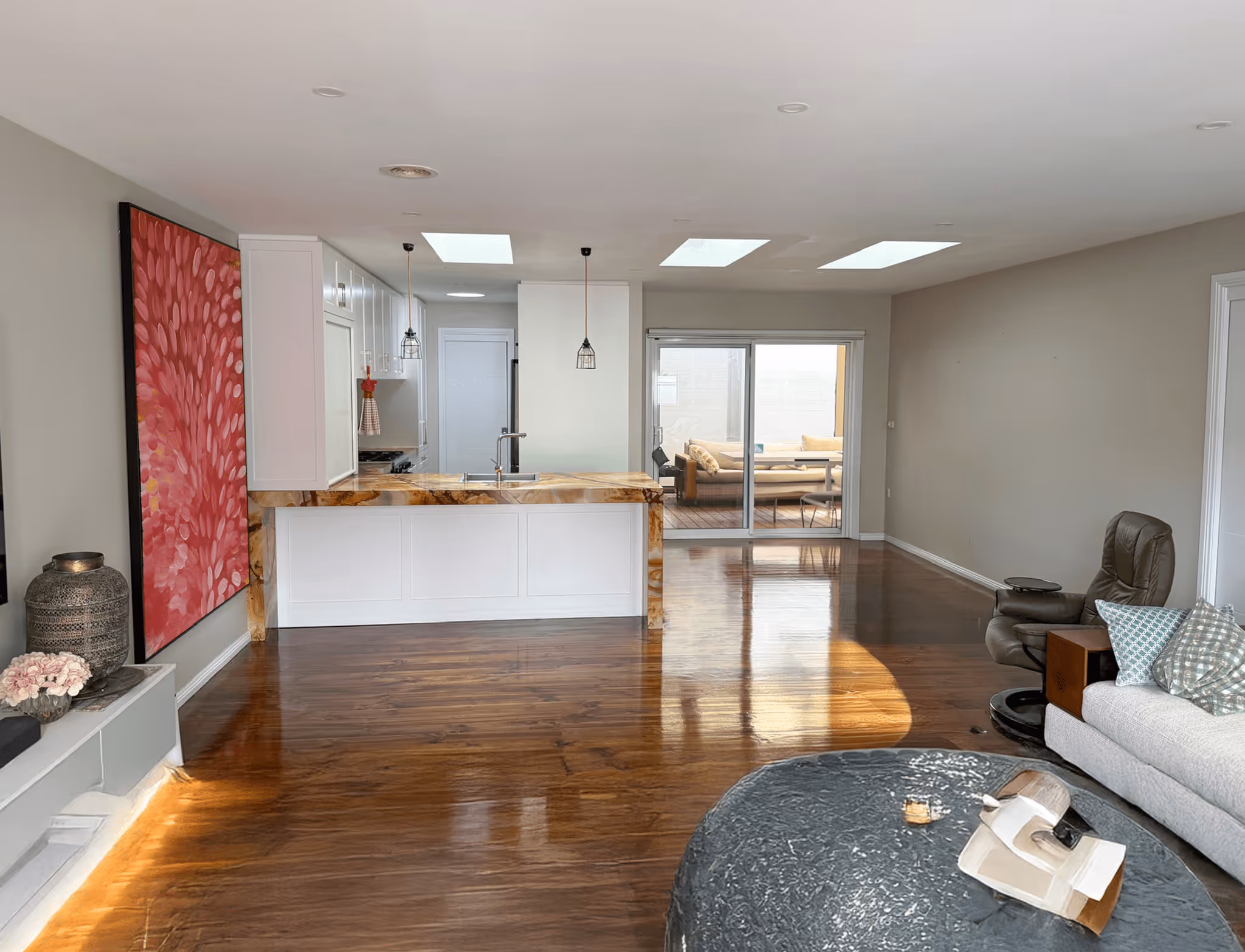Spacious modern living area with polished wooden floor, kitchen island with marble countertop, large red floral painting, and sliding glass door leading to outdoor seating.