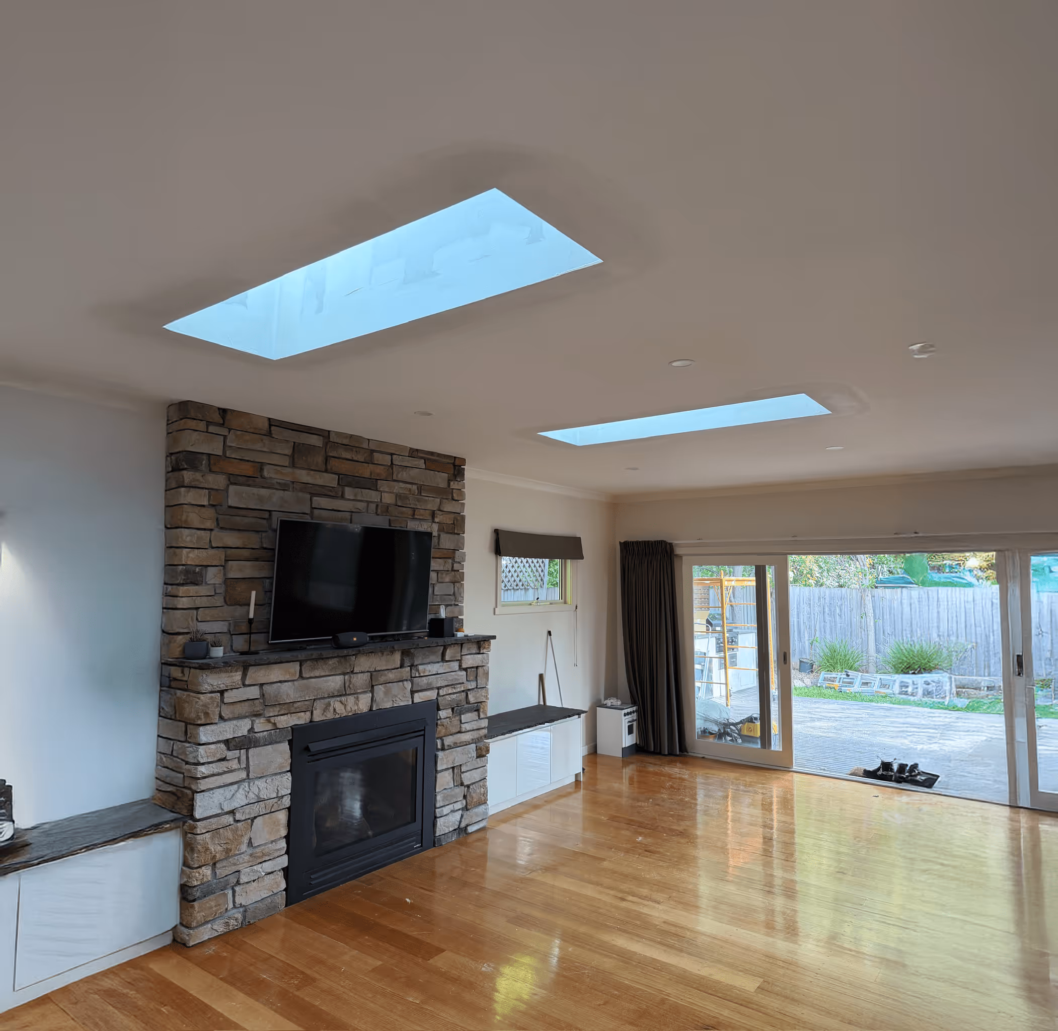 Living room with hardwood floor, stone fireplace with mounted TV, two skylights, and large sliding glass doors overlooking a fenced backyard.