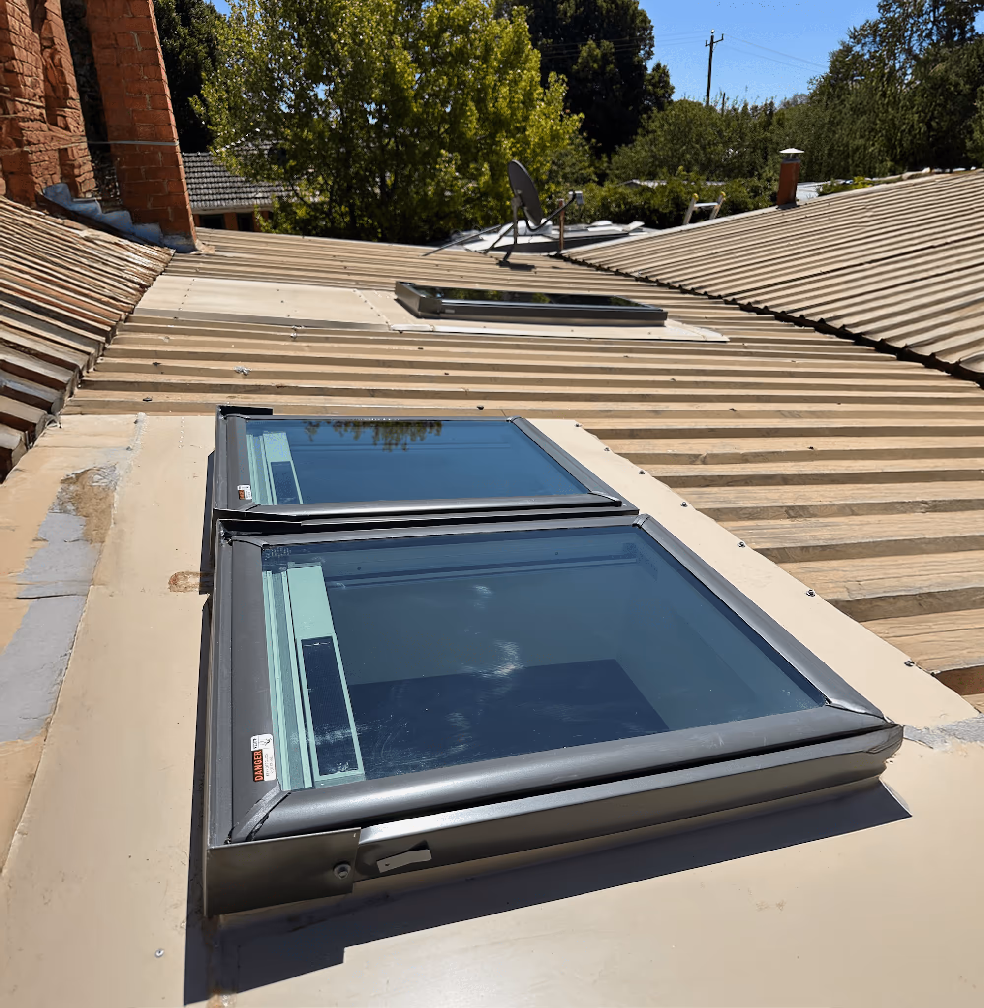 Two skylight windows installed on a metal roof with a satellite dish and green trees in the background under a clear blue sky.