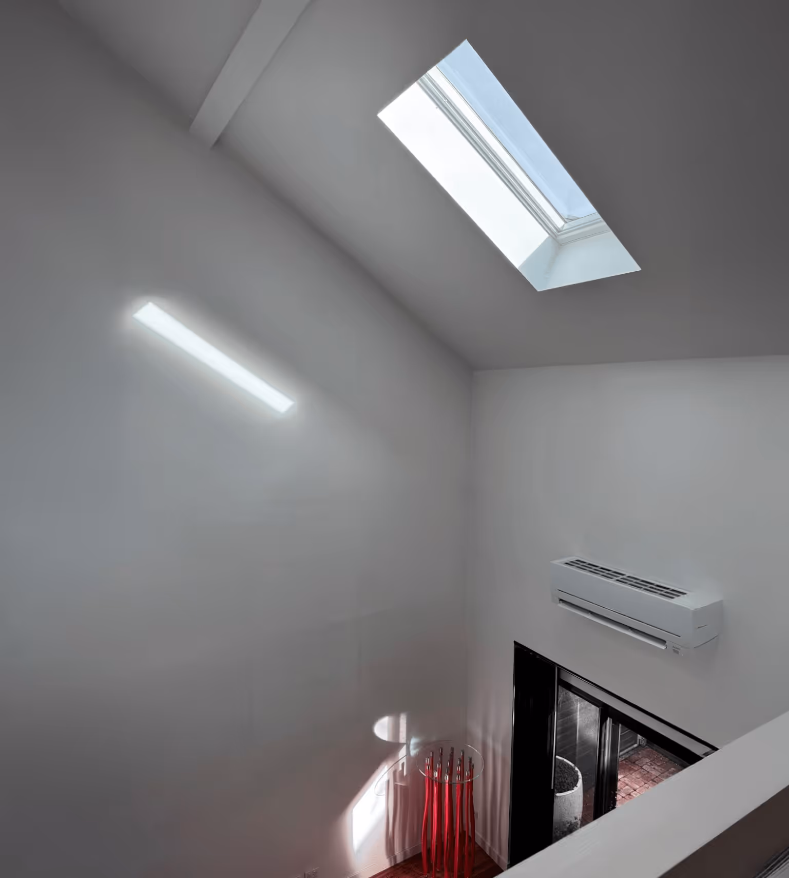 Interior of a bright room with a skylight, wall-mounted air conditioner, glass door leading outside, and a modern red umbrella stand casting shadows.
