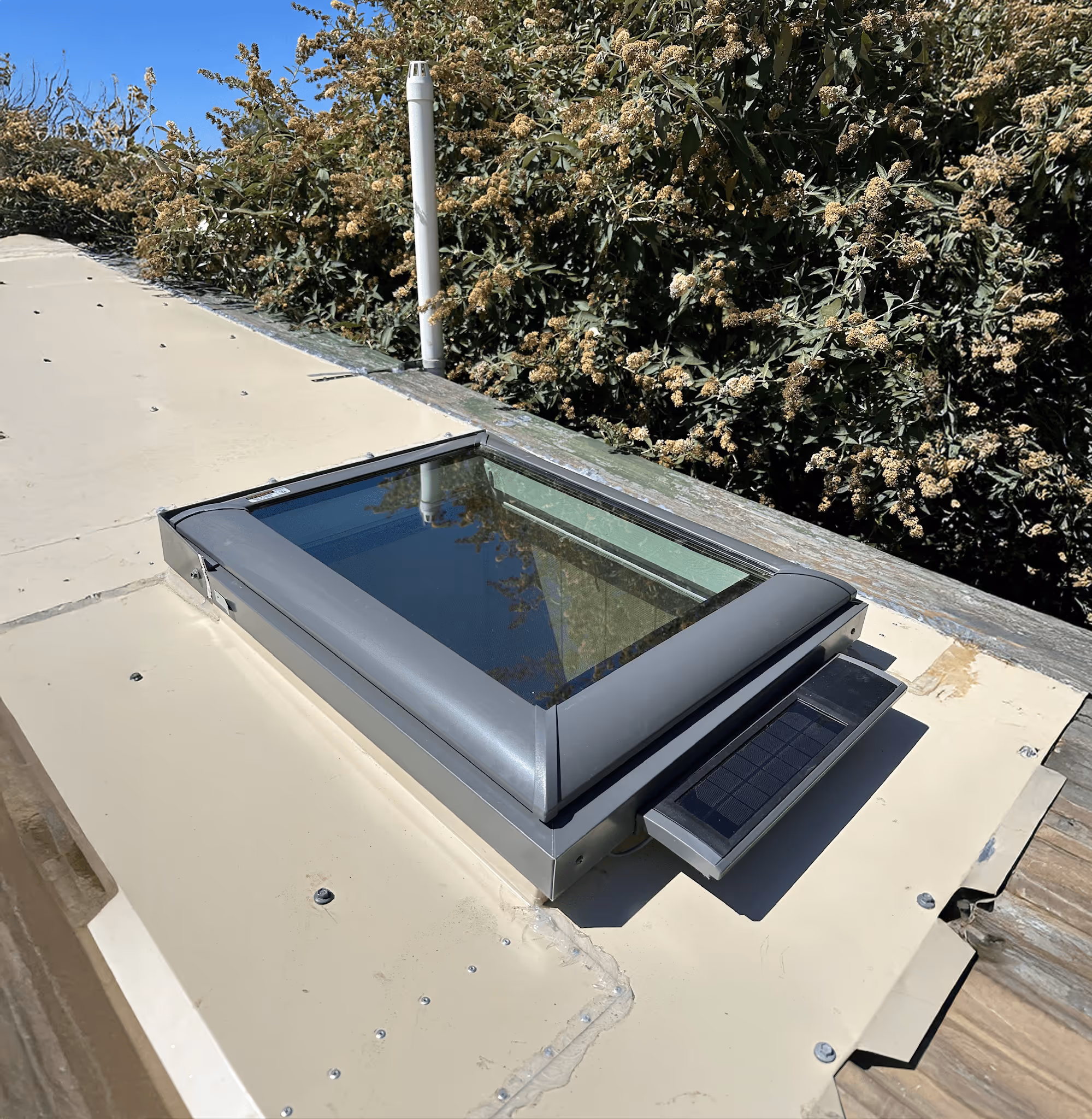 Rectangular roof window with a solar panel on one side installed on a beige flat roof under a clear blue sky.