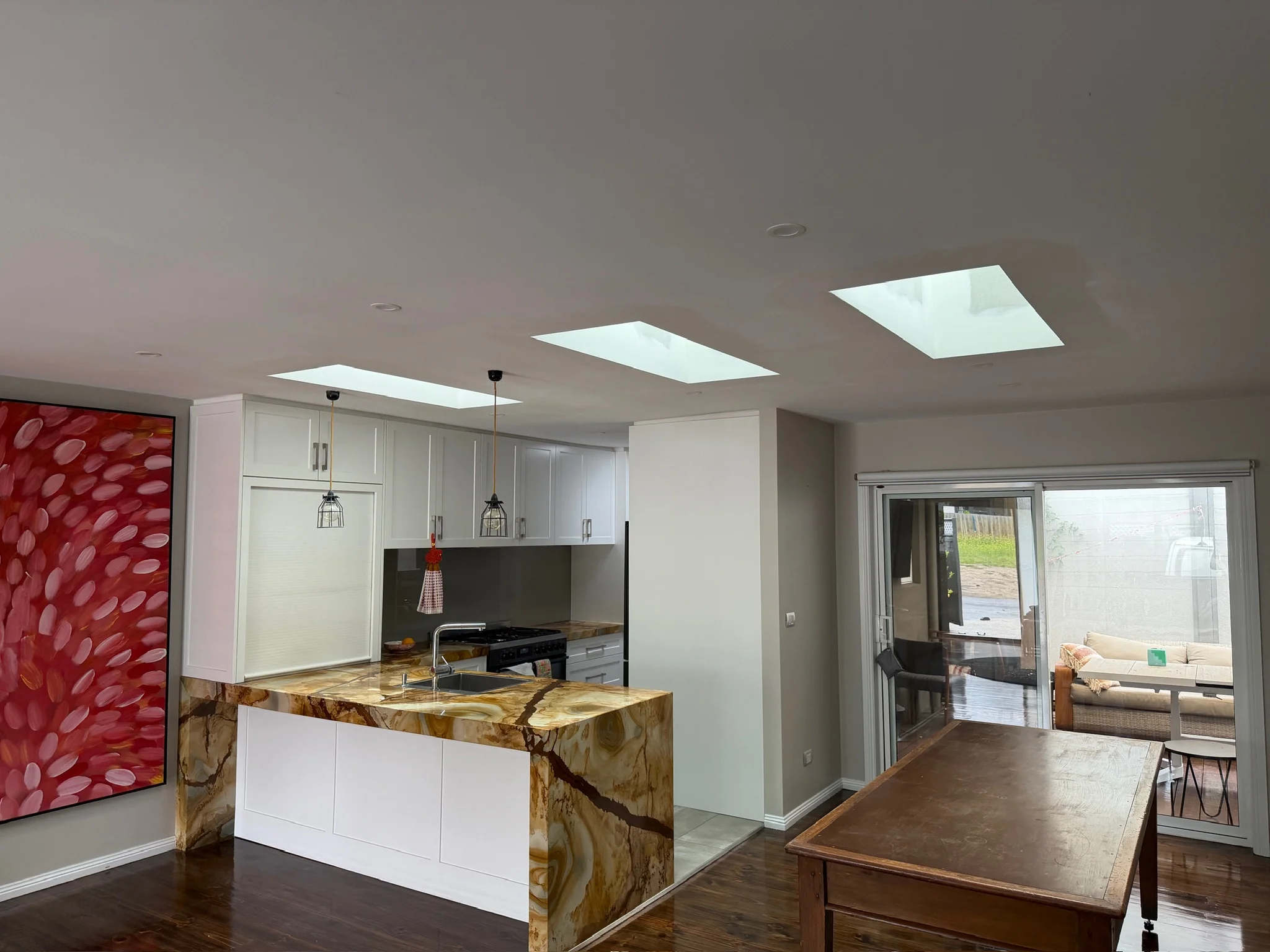 Modern open kitchen with white cabinets, marble countertop, two pendant lights, hardwood floor, and adjacent living area with couch and armchair.