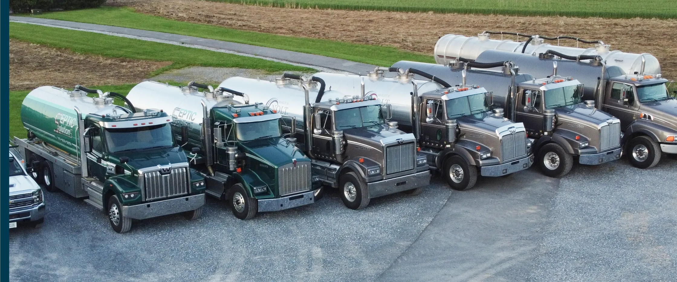 Lineup of various large trucks including septic tankers, a white pickup with excavator, and heavy-duty semi trucks parked on gravel.