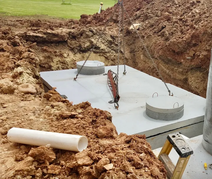Concrete septic tank being installed in an excavated hole with surrounding soil and a white PVC pipe nearby.