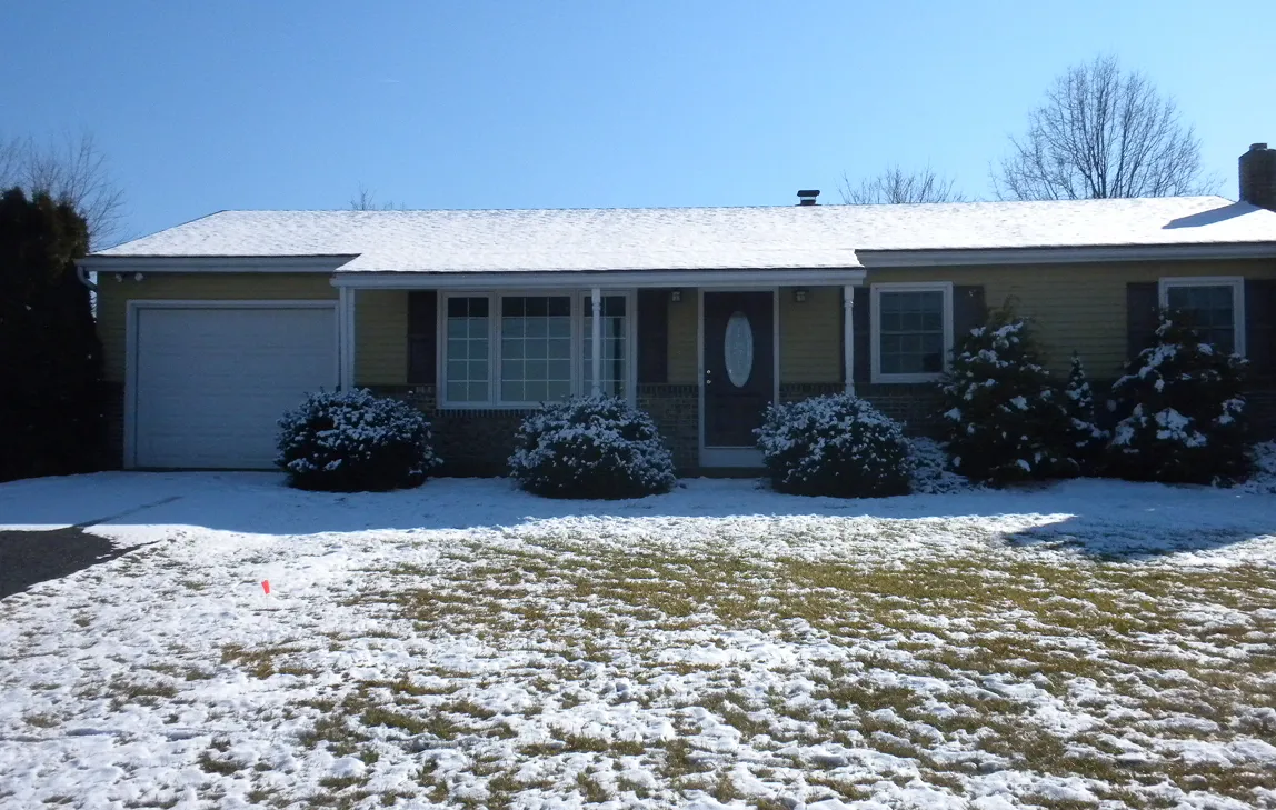 Single-story house with a snow-covered roof and partially snow-covered lawn under a clear blue sky.