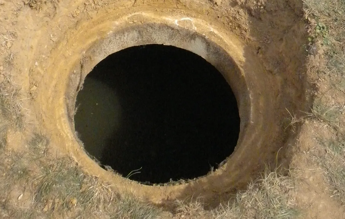 Open circular hole in dry soil with some grass around the edges.