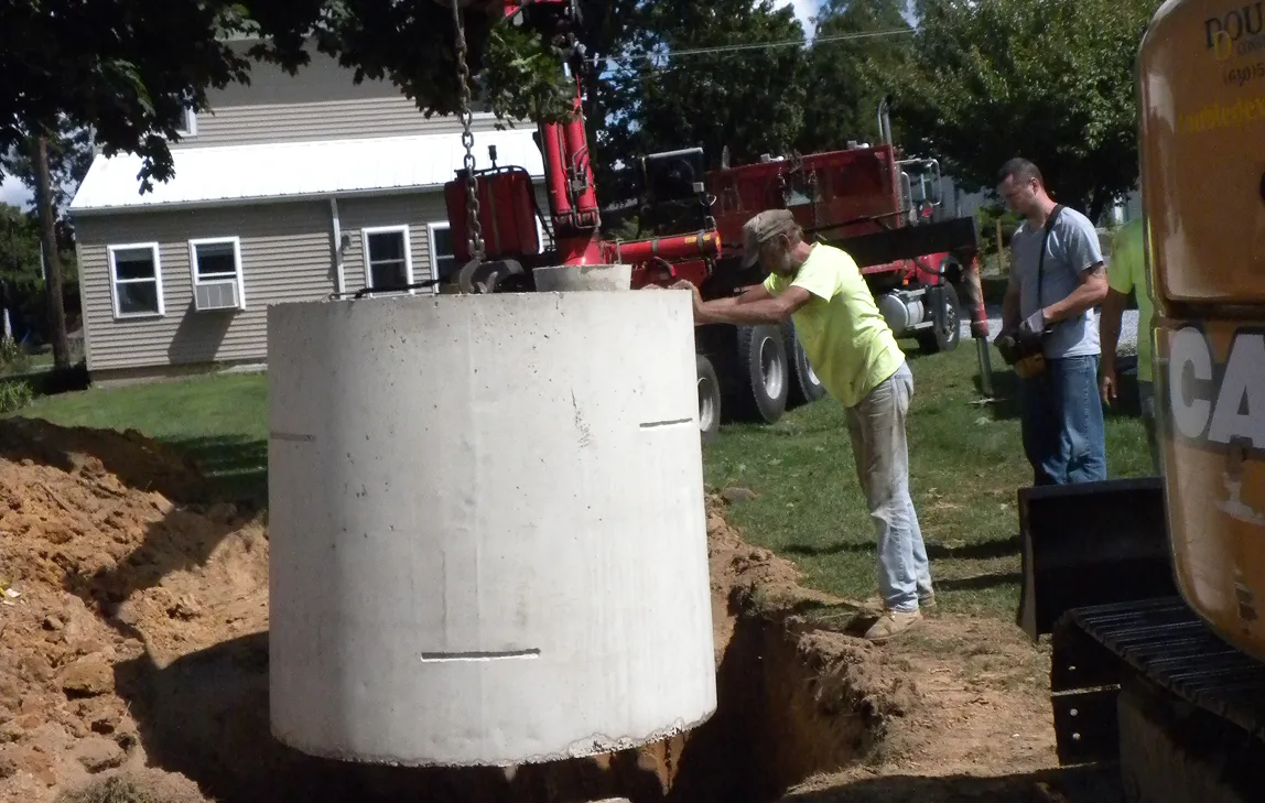 Two construction workers guiding a large concrete cylinder being lowered into a deep dug hole in a residential yard.