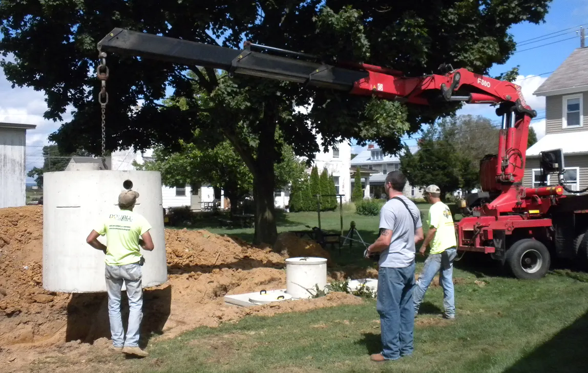 Three construction workers guiding a large concrete pipe being lifted by a crane truck at an excavation site in a residential neighborhood.