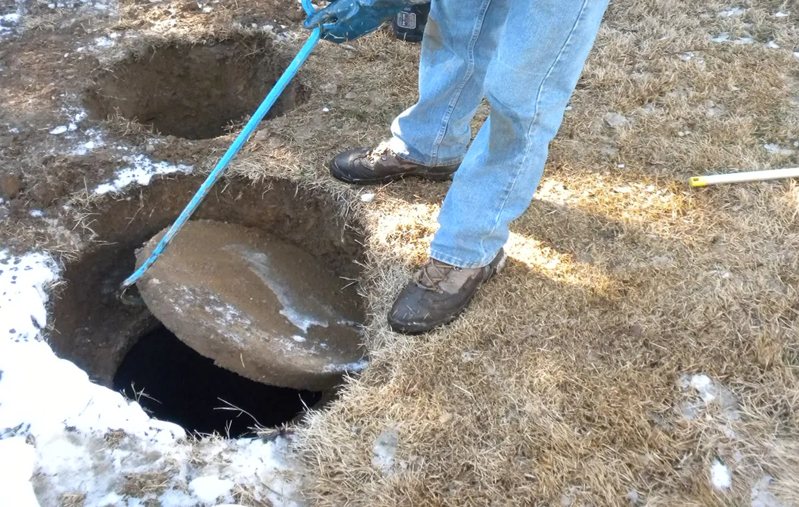 Worker wearing jeans and boots lifting a round concrete manhole cover with a blue metal hook near a hole in dry grass partially covered with patches of snow.