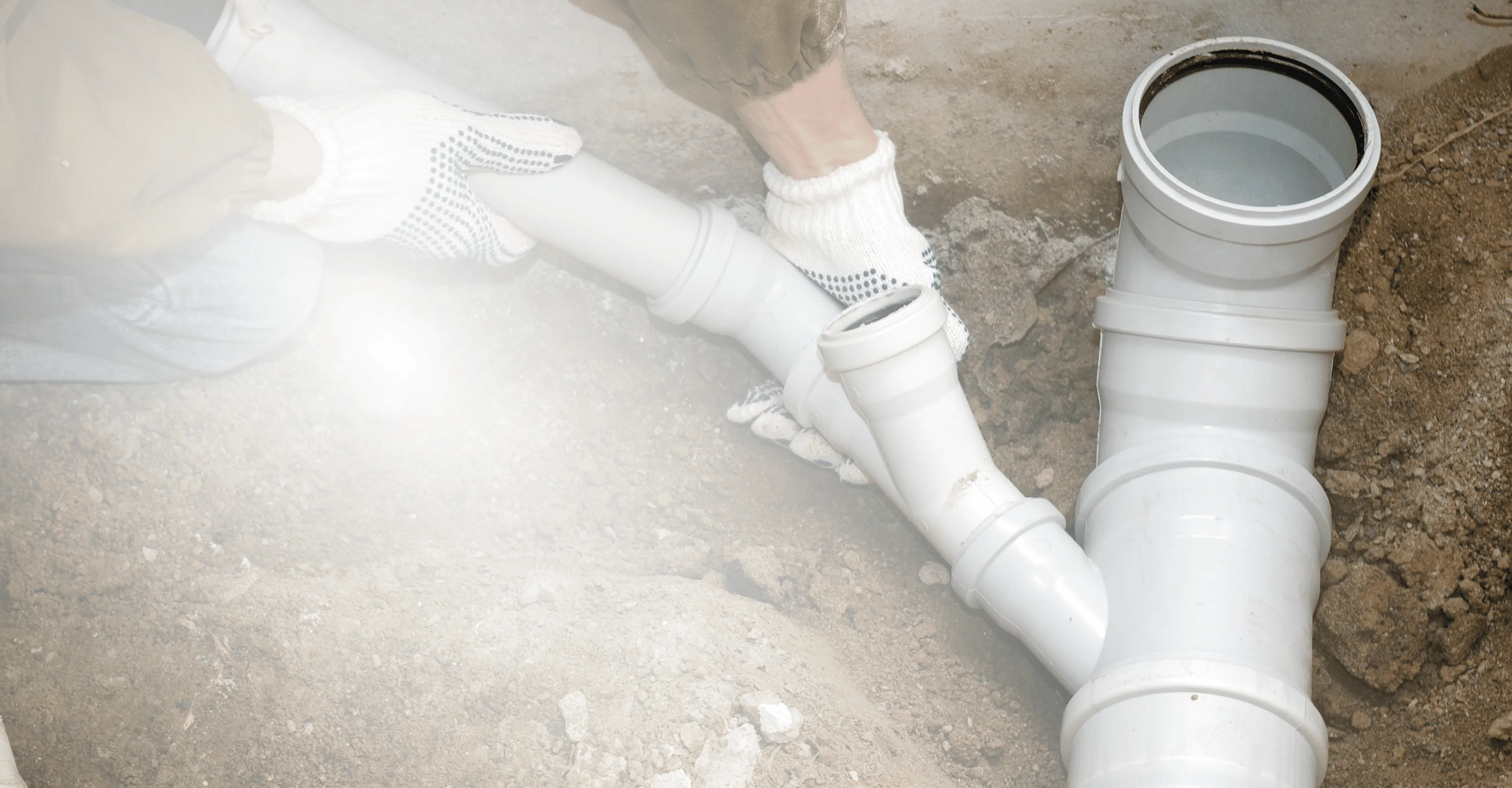 A worker wearing white gloves assembling white PVC pipes in the ground for plumbing.