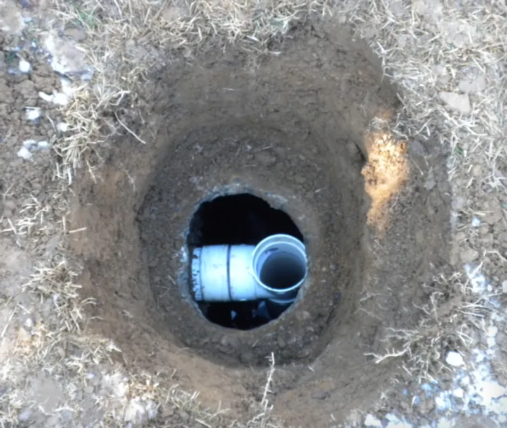 A small dug hole in dry soil with a visible white PVC pipe inside.