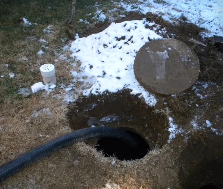 Open hole in a grassy area partially covered with snow, with a large hose inserted into it and a round concrete cover nearby.