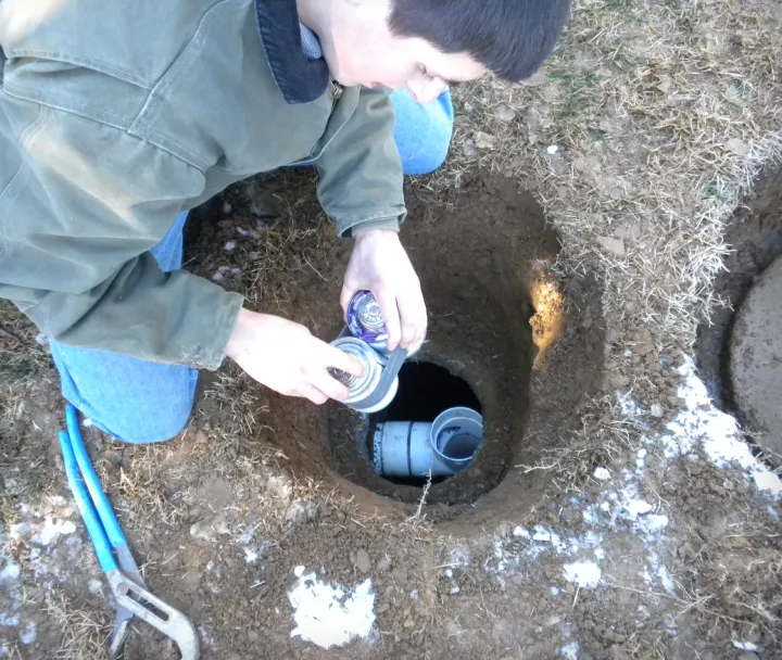 Person kneeling beside a round hole in the ground, holding two cans over a pipe inside the hole with snow patches around.