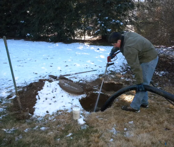 Person using a water jet tool to clean or unblock a pipe outdoors in a snowy yard.