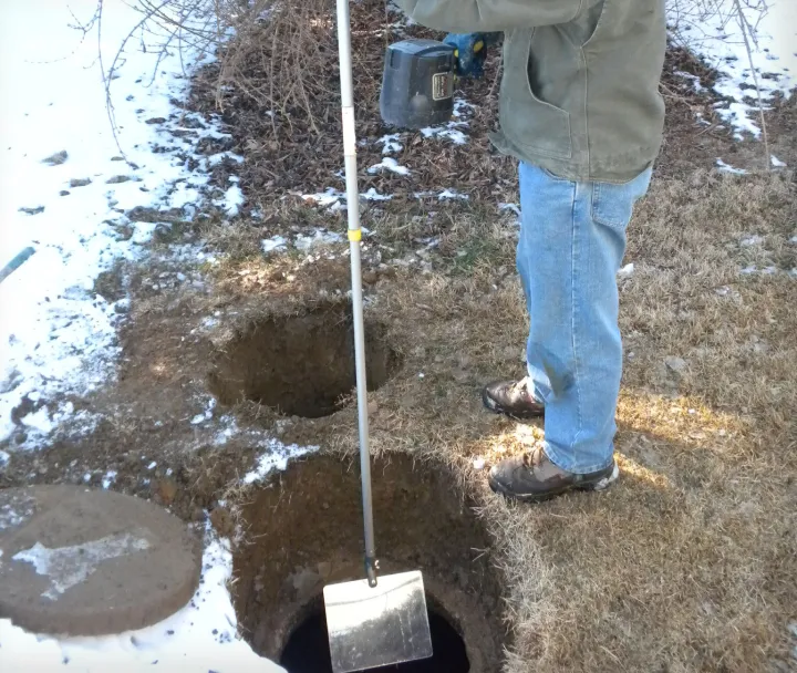 Person standing next to two round holes in the ground, one with a metal shovel-like tool inserted, surrounded by patches of snow and dry grass.