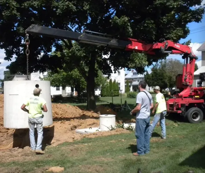 Three construction workers guiding a large concrete pipe being lifted by a crane truck at an excavation site in a residential neighborhood.